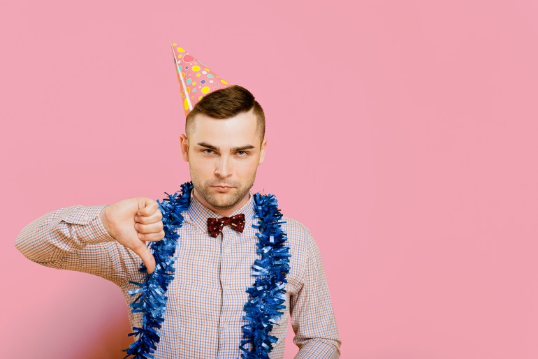 Young adult guy 20-25s wearing a party hat and New Year's tinsel, unhappy and angry showing a thumbs down or dislike gesture, frustrated with a displeased expression.